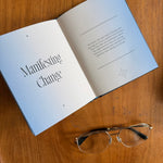 An open book titled 'Manifesting Change' lies on a wooden table, with a pair of metal-framed eyeglasses placed below it.
