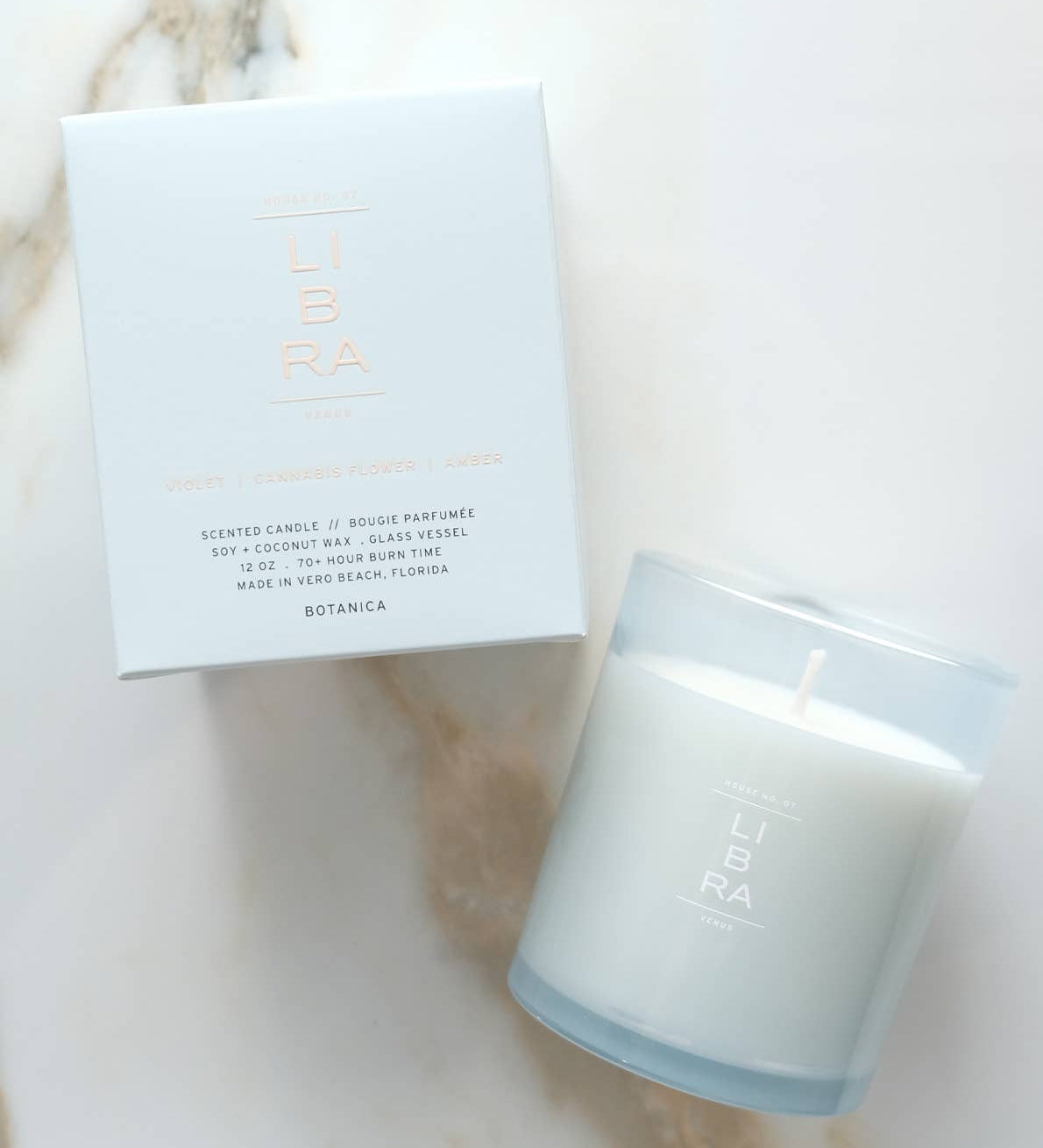 A white scented candle in a frosted glass jar next to its white packaging box, both featuring the brand name 'LIBRA' in a metallic rose-gold tone, set on a light marble surface.