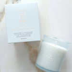 A white scented candle in a frosted glass jar next to its white packaging box, both featuring the brand name 'LIBRA' in a metallic rose-gold tone, set on a light marble surface.