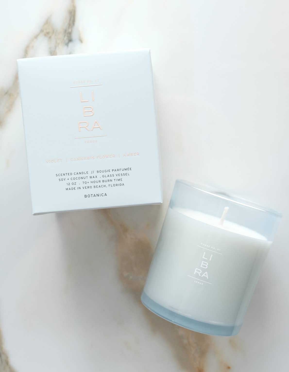 A white scented candle in a frosted glass jar next to its white packaging box, both featuring the brand name 'LIBRA' in a metallic rose-gold tone, set on a light marble surface.