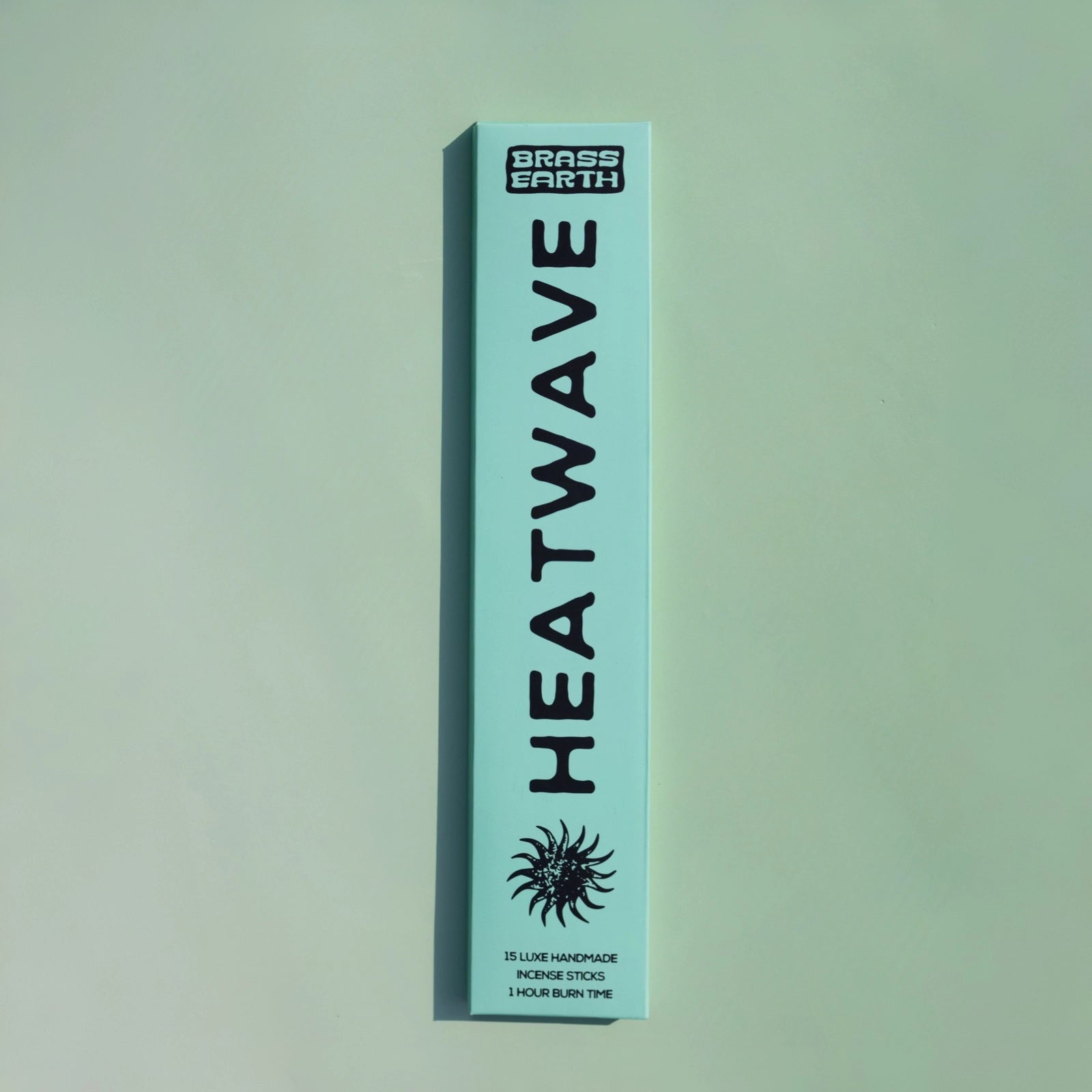 A tall, narrow mint-green box for Grass Earth Heatwave incense sticks, featuring bold black text and a sun graphic, set against a matching mint-green background.