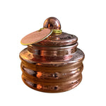 A decorative copper-colored lidded jar with a ribbed base and a small round knob on top, shown on a plain white background. A round paper tag is attached to the lid and appears to read 'Paddywax Pink Peppermint' as the visible brand.