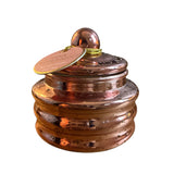 A decorative copper-colored lidded jar with a ribbed base and a small round knob on top, shown on a plain white background. A round paper tag is attached to the lid and appears to read 'Paddywax Pink Peppermint' as the visible brand.