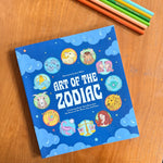 The book 'Art of the Zodiac' by Jess Miller is displayed on a wooden table, surrounded by colored pencils. The cover is bright blue with illustrations of various zodiac signs in circular frames and white title text.