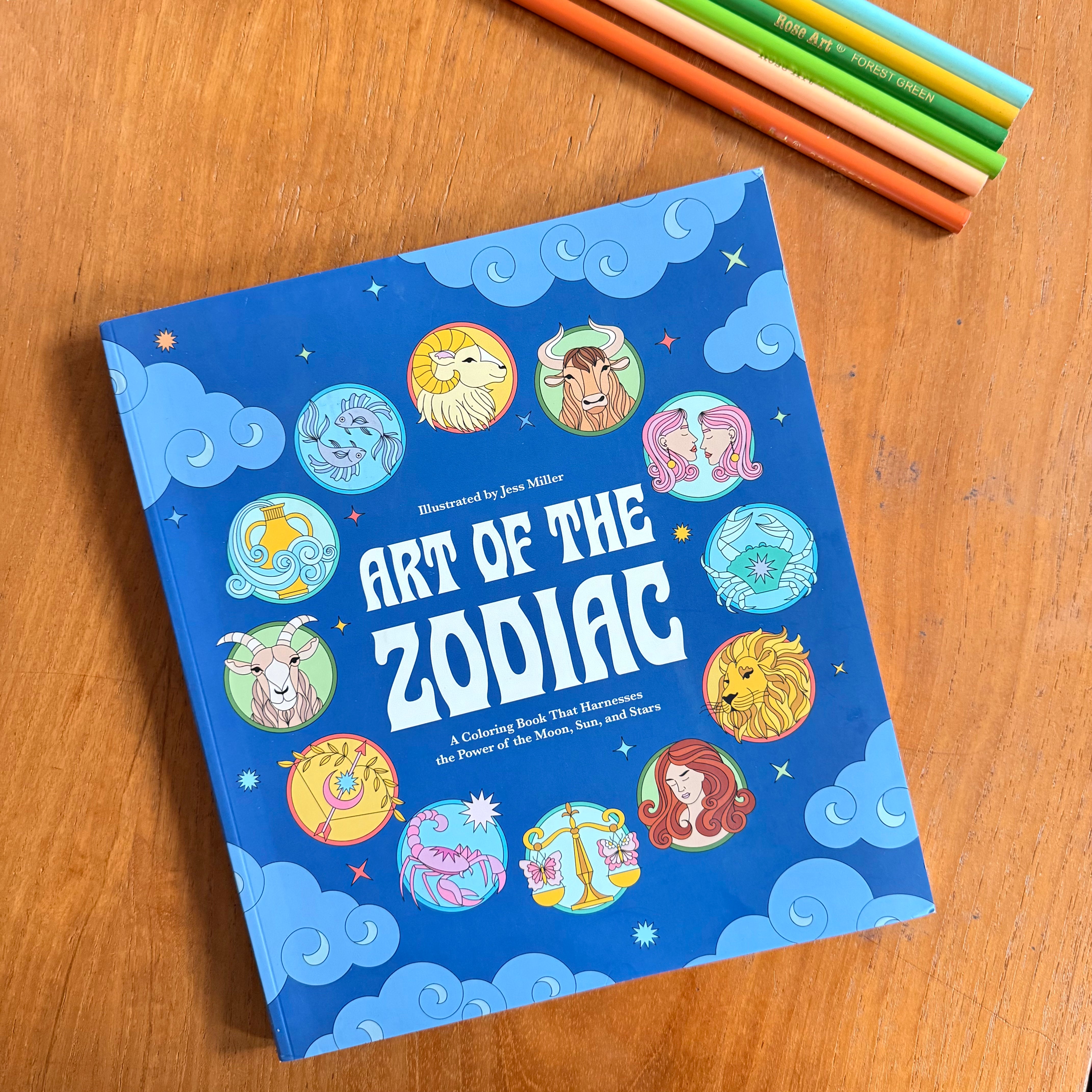 The book 'Art of the Zodiac' by Jess Miller is displayed on a wooden table, surrounded by colored pencils. The cover is bright blue with illustrations of various zodiac signs in circular frames and white title text.