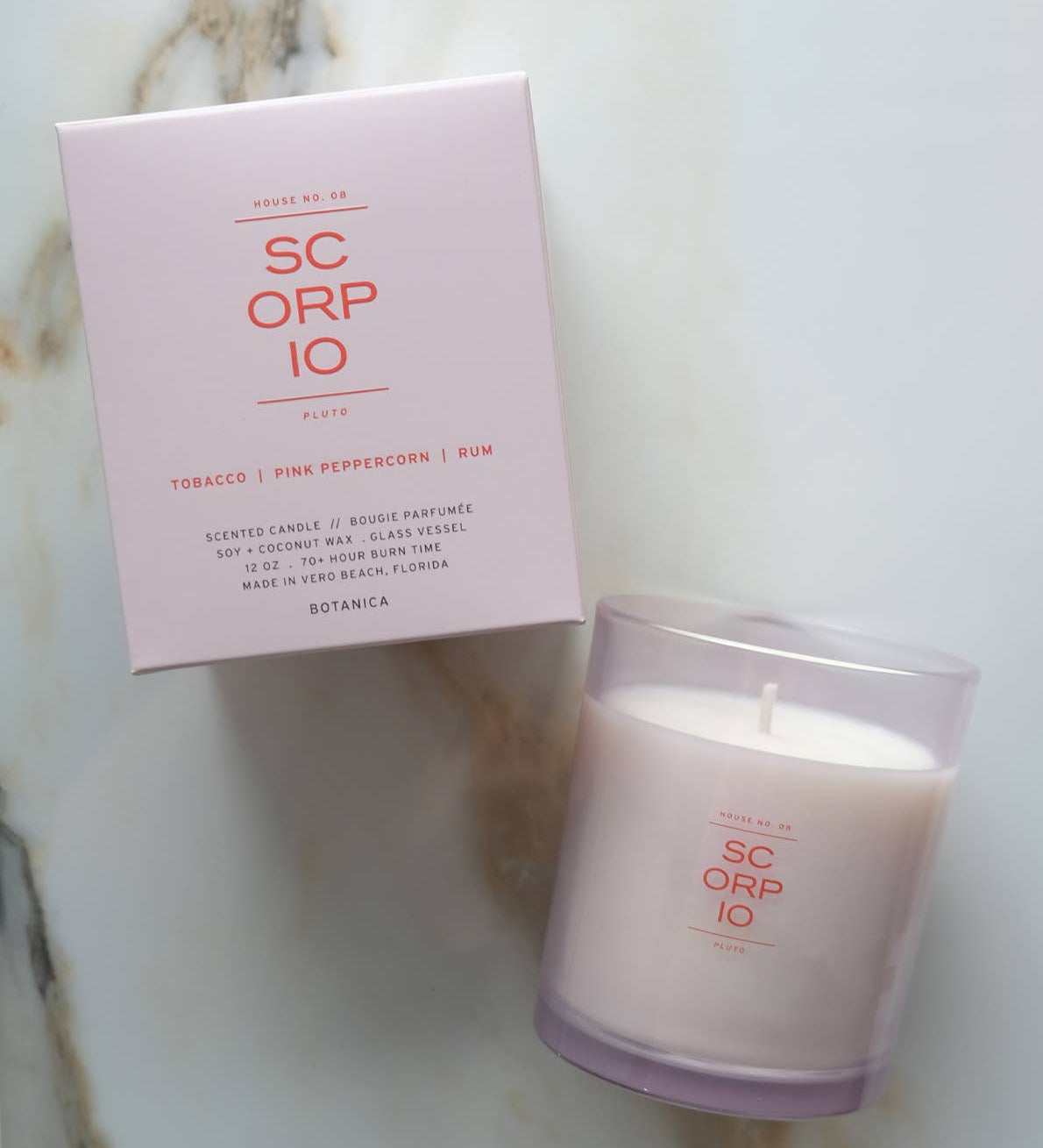 A SC ORP IO scented candle in a frosted glass jar next to its light pink packaging box, placed on a white marble surface.