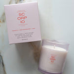 A SC ORP IO scented candle in a frosted glass jar next to its light pink packaging box, placed on a white marble surface.