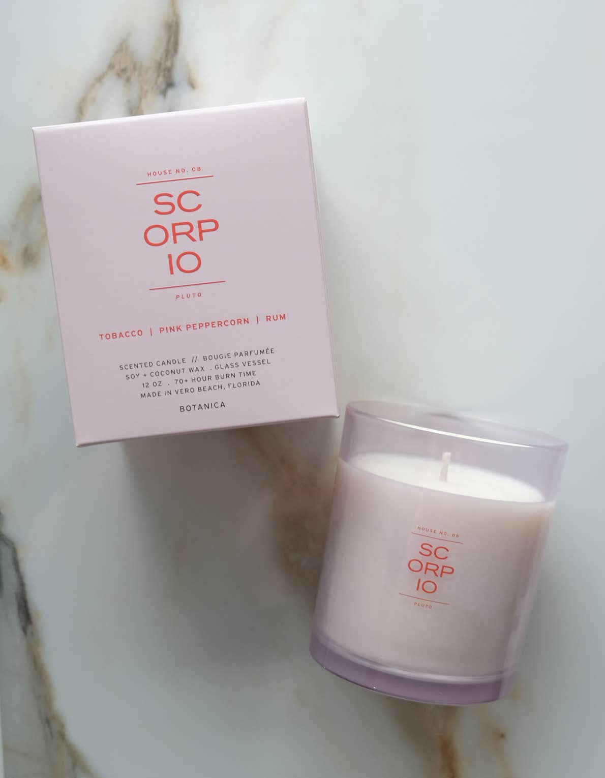 A SC ORP IO scented candle in a frosted glass jar next to its light pink packaging box, placed on a white marble surface.