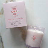 A SC ORP IO scented candle in a frosted glass jar next to its light pink packaging box, placed on a white marble surface.