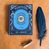 A hardcover book titled 'Manifesting: Live and Dream Life' by Greta Ellin, placed on a wooden surface next to a black feather and a small wooden tumbler.
