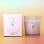 A Botanica 'Taurus' scented candle in a clear glass jar next to its light pink packaging box, displayed on warm orange gradient background