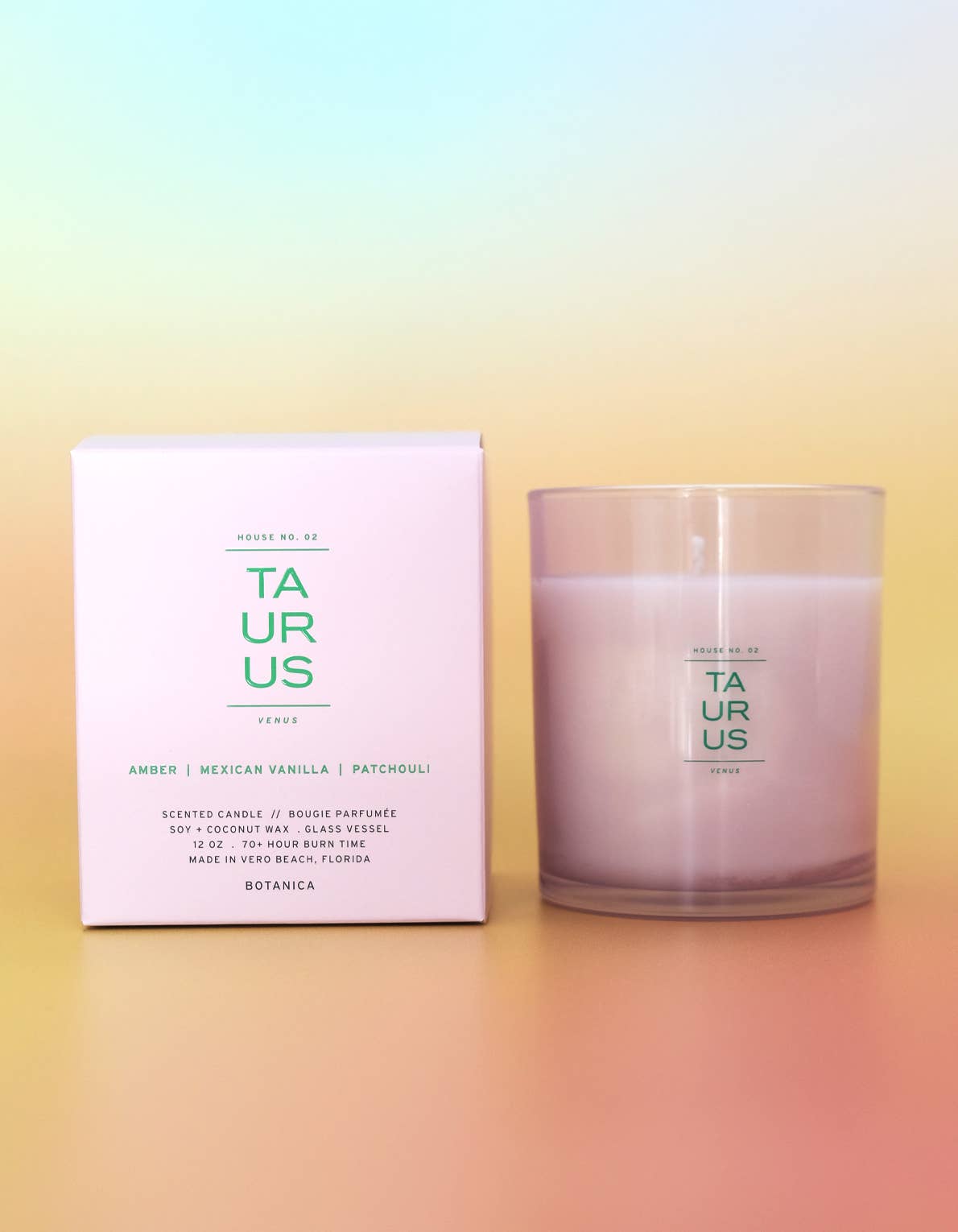 A Botanica 'Taurus' scented candle in a clear glass jar next to its light pink packaging box, displayed on warm orange gradient background