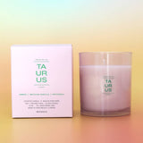 A Botanica 'Taurus' scented candle in a clear glass jar next to its light pink packaging box, displayed on warm orange gradient background