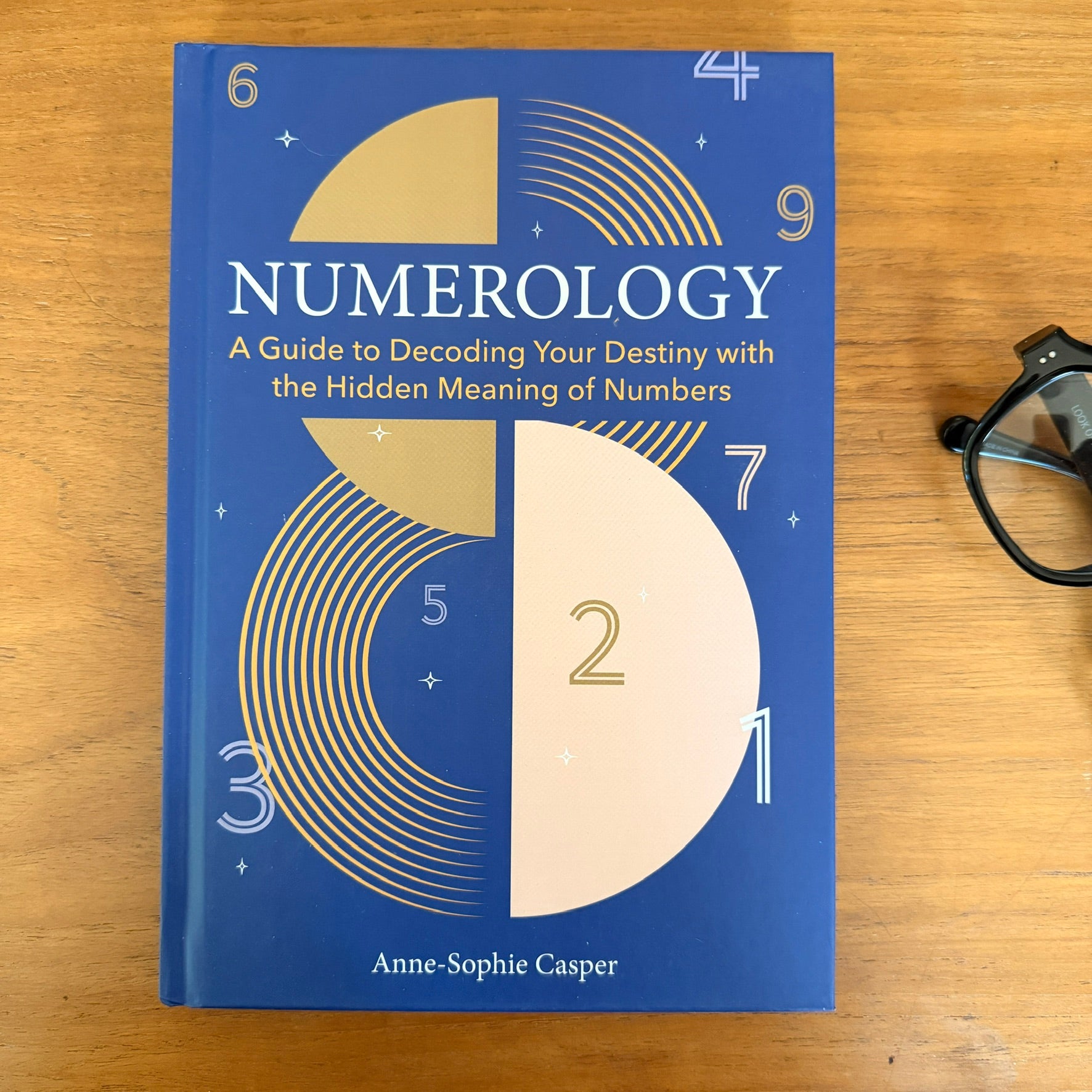 Blue book cover with Numerology: A Guide to Decoding Your Destiny with the Hidden Meaning of Numbers on a wood background with a pair of black reading glasses
