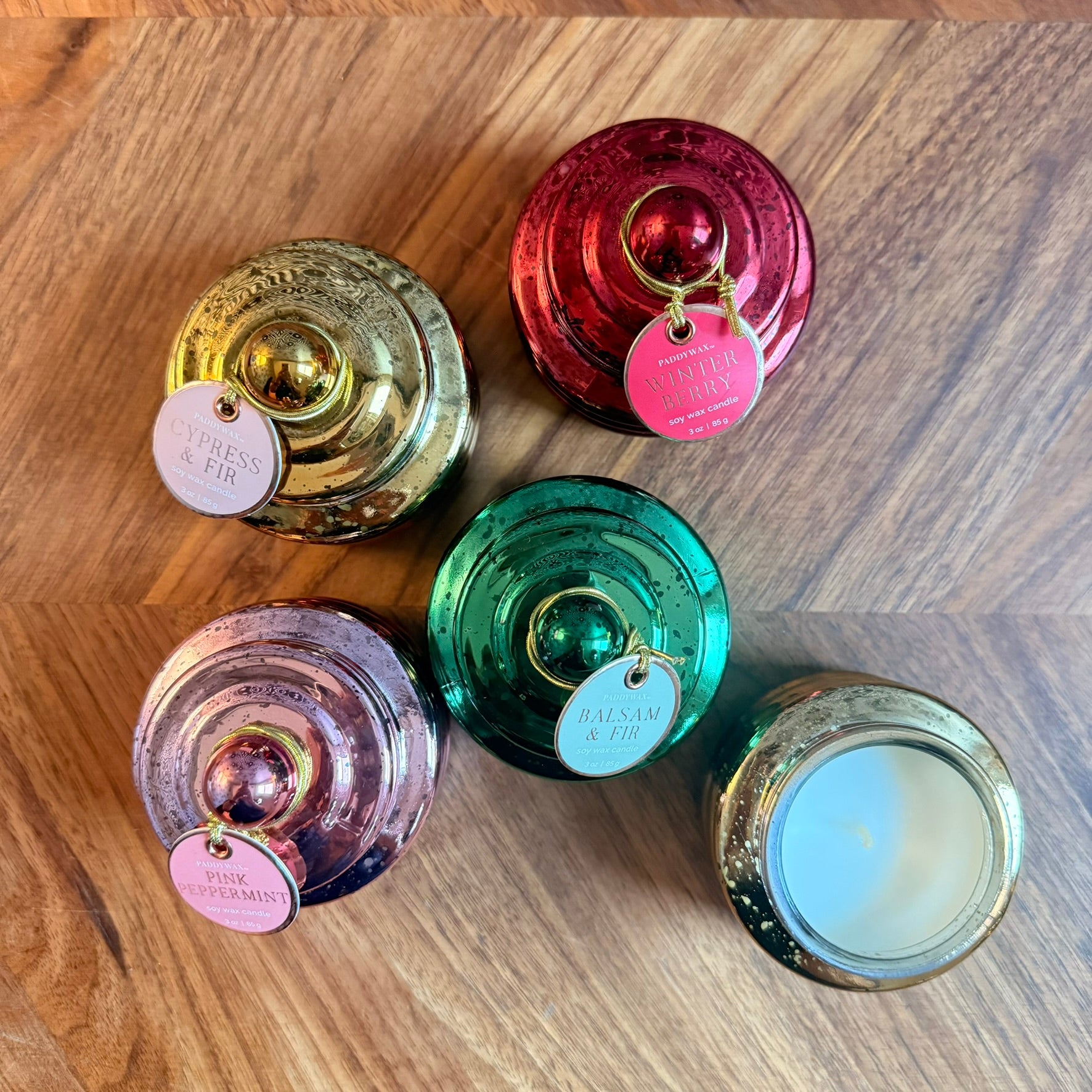 5 round shiny vessels with Paddywax Cypress & Fir, Pink Peppermint, Balsam & Fir and WinterBerry labels tied on and a gold round candle on a wood tabletop