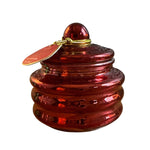 A small round amber-red glass lidded container with a ribbed base, topped with a matching glass knob and a gold-tone wire loop holding a small red tag, shown on a plain white background.