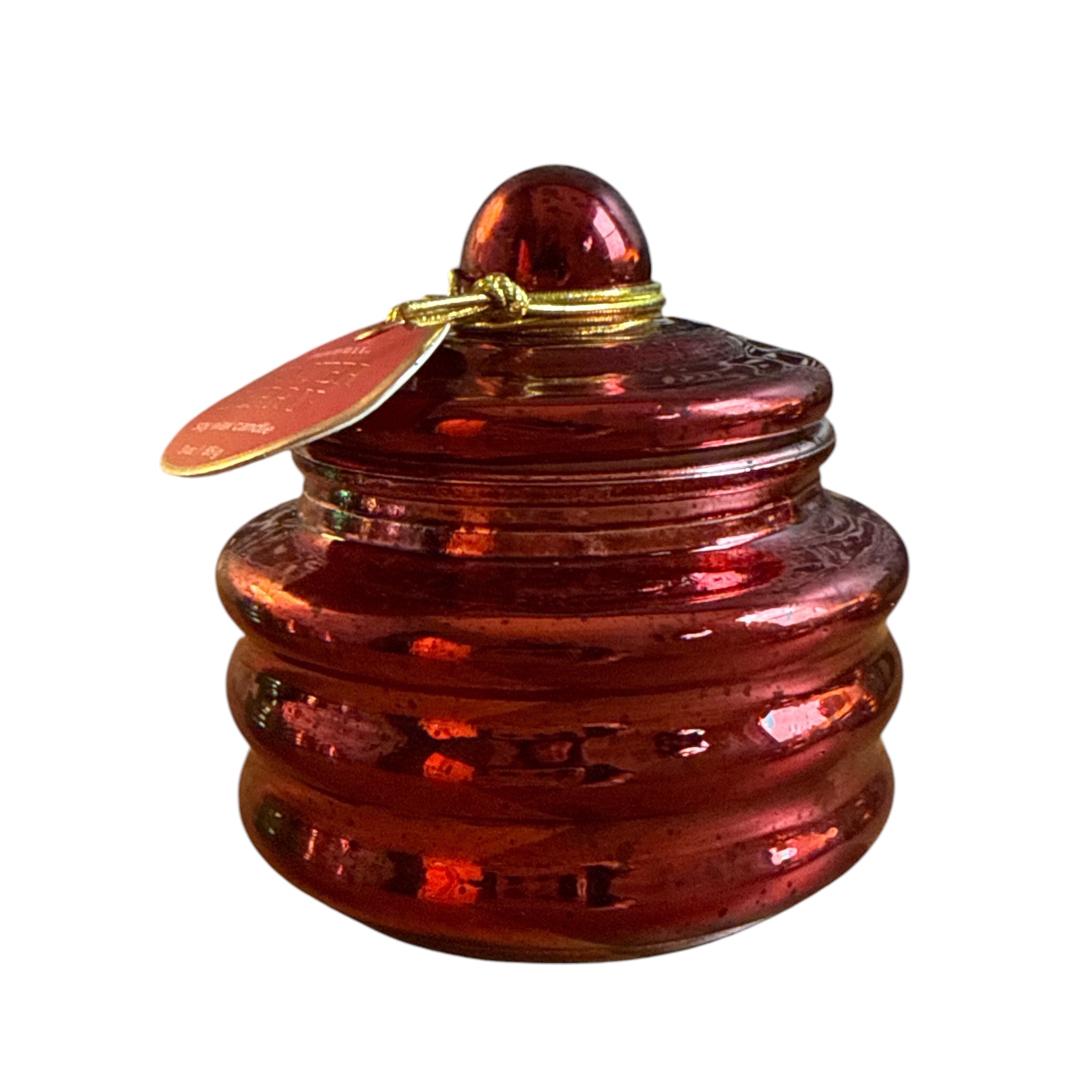 A small round amber-red glass lidded container with a ribbed base, topped with a matching glass knob and a gold-tone wire loop holding a small red tag, shown on a plain white background.