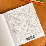 A black and white line art coloring page featuring a Virgo woman surrounded by plants, shown on a wooden table with colored pencils in the background.