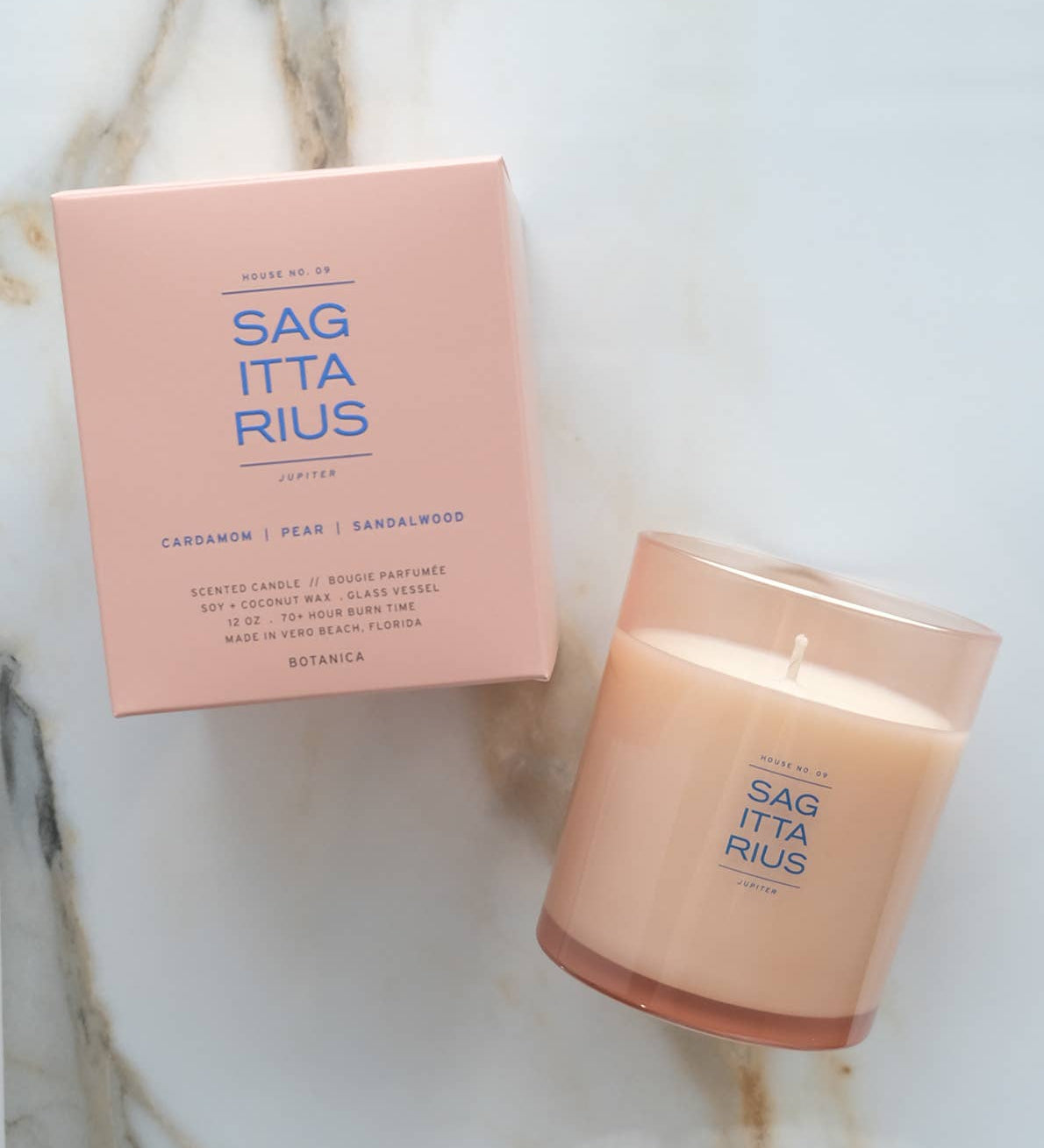 A lit candle in a frosted glass jar next to its matching pink packaging box, both featuring the brand name 'SAG ITTA RIUS' on a light marble surface.