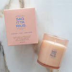 A lit candle in a frosted glass jar next to its matching pink packaging box, both featuring the brand name 'SAG ITTA RIUS' on a light marble surface.