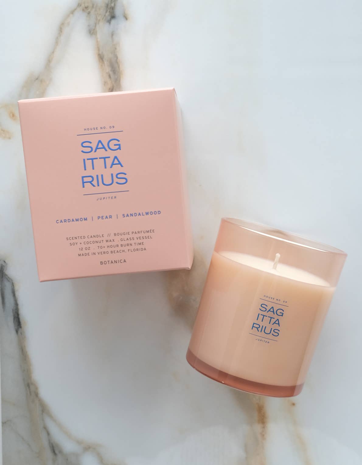 A lit candle in a frosted glass jar next to its matching pink packaging box, both featuring the brand name 'SAG ITTA RIUS' on a light marble surface.