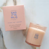 A lit candle in a frosted glass jar next to its matching pink packaging box, both featuring the brand name 'SAG ITTA RIUS' on a light marble surface.