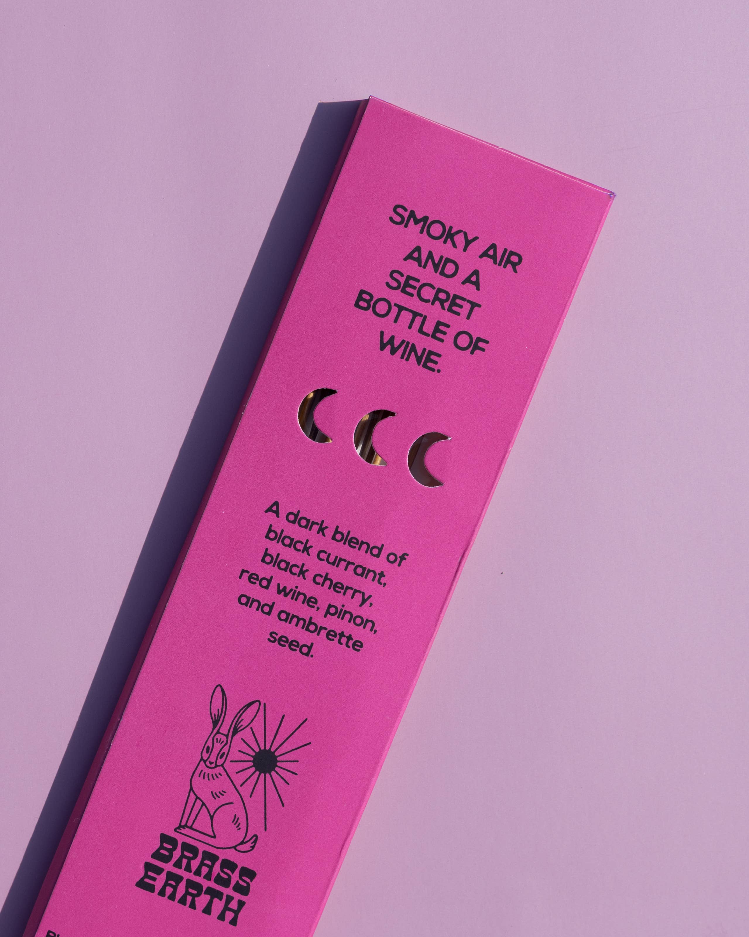 Bright pink rectangular product packaging for Brass Earth wine, photographed at an angle on a matching pink background. The box features black text reading 'SMOKY AIR AND A SECRET BOTTLE OF WINE.