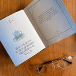 An open book or journal laid on a wooden surface, showing two interior pages with printed text and a small teapot illustration. A pair of gold-framed eyeglasses rests on the lower right corner of the page.