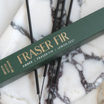 A dark green incense box labeled 'FRASER FIR' with gold lettering, placed diagonally over several thin black incense sticks on a white and gray marbled surface.