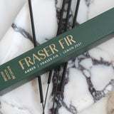 A dark green incense box labeled 'FRASER FIR' with gold lettering, placed diagonally over several thin black incense sticks on a white and gray marbled surface.