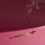 Incense stick burning on a brass incense holder with smoke against a pink background