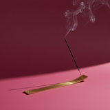 Incense stick burning on a brass incense holder with smoke against a pink background