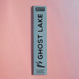 A tall, narrow light blue box for Brass Earth Ghost Lake incense sticks, standing vertically against a soft pink gradient background.