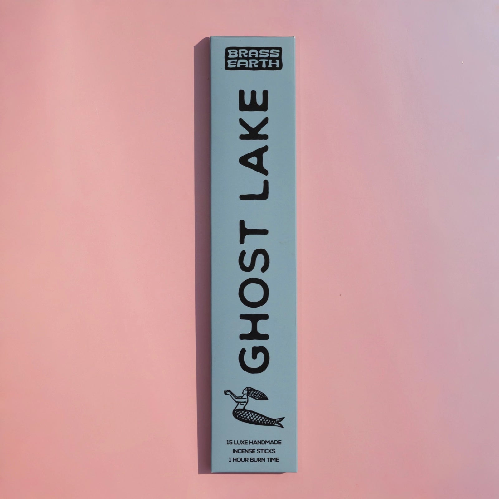 A tall, narrow light blue box for Brass Earth Ghost Lake incense sticks, standing vertically against a soft pink gradient background.