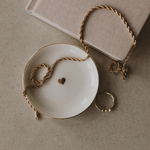 Necklace  on a white dish with a gold heart, next to a book and ring on a beige surface