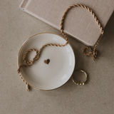 Necklace  on a white dish with a gold heart, next to a book and ring on a beige surface