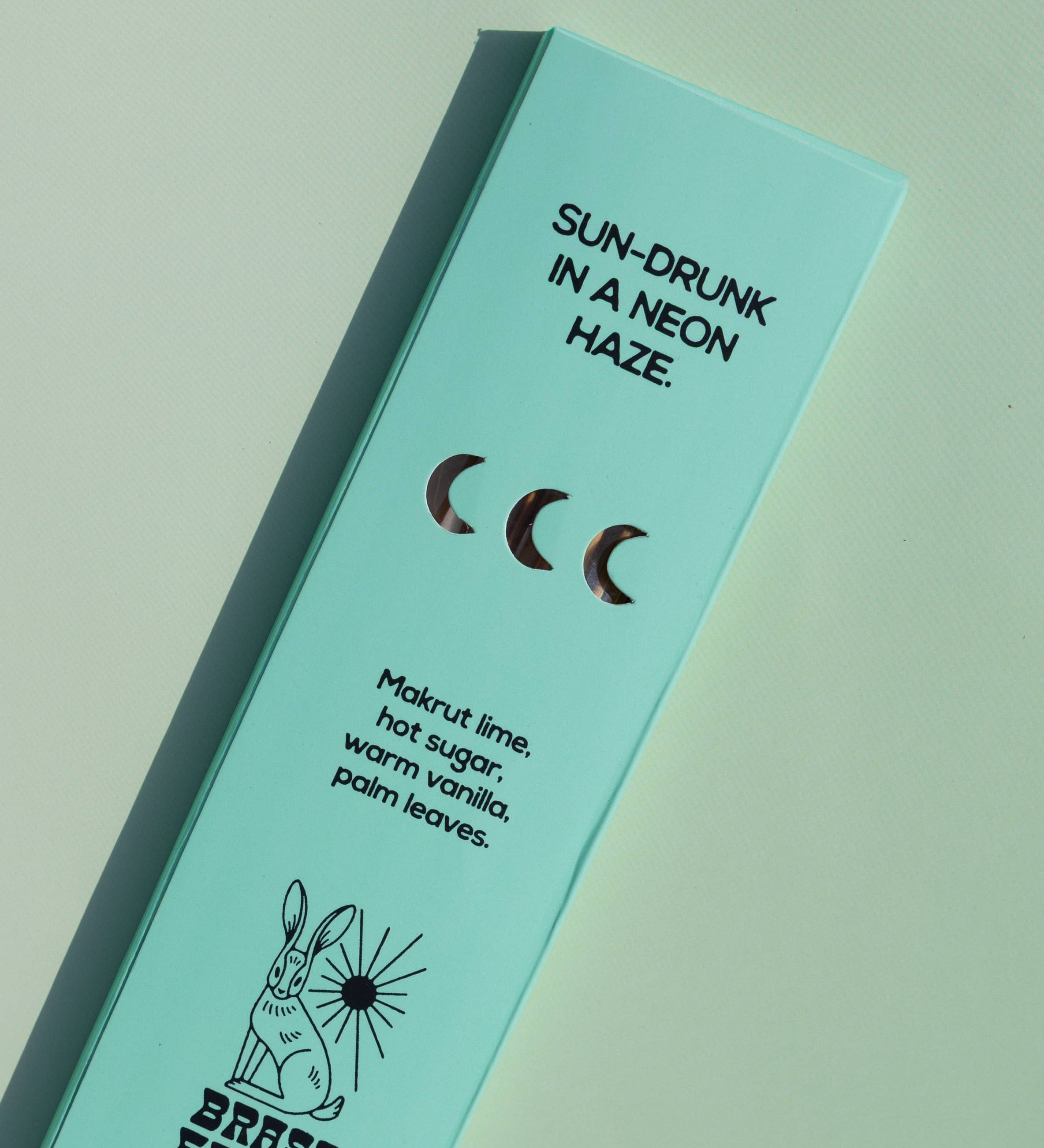 A mint green rectangular product box for Brass Earth, photographed at an angle on a matching mint background. The box features black text reading 'SUN-DRUNK IN A NEON HAZE.