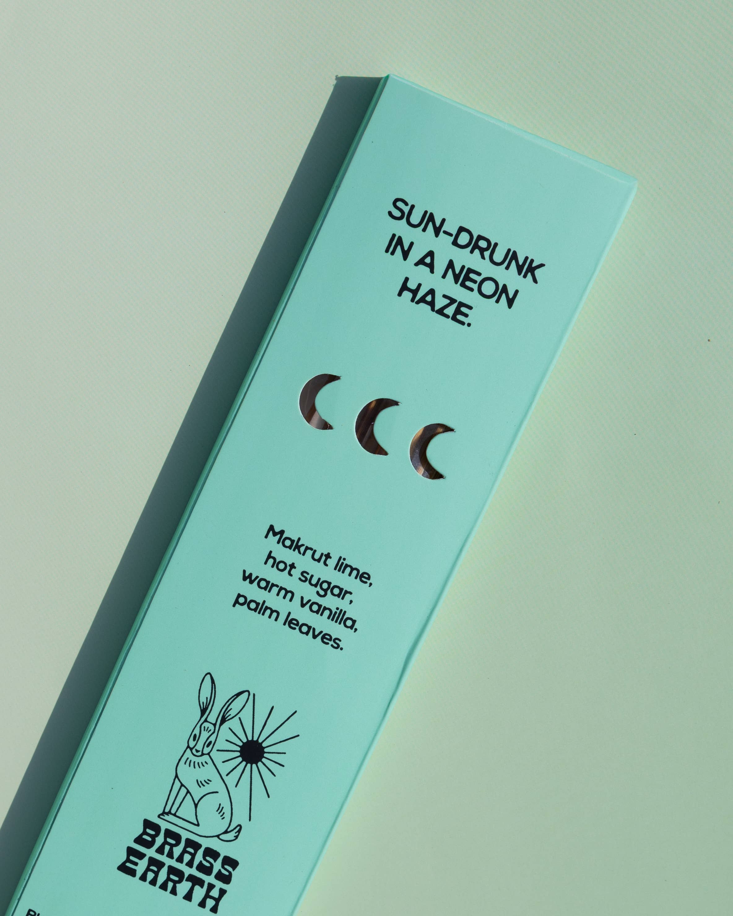 A mint green rectangular product box for Brass Earth, photographed at an angle on a matching mint background. The box features black text reading 'SUN-DRUNK IN A NEON HAZE.