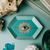 Teal decorative dish with eye design on a teal surface with rocks, books, and a bust.