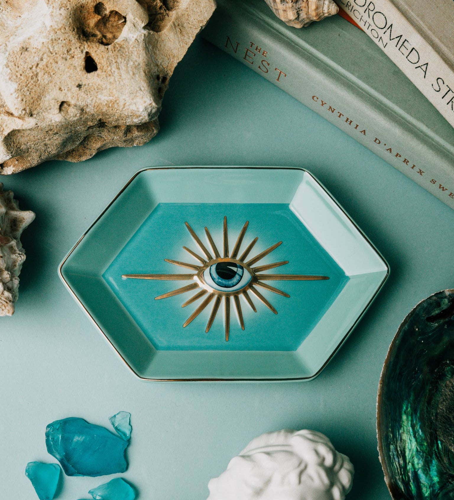 Teal decorative dish with eye design on a teal surface with rocks, books, and a bust.