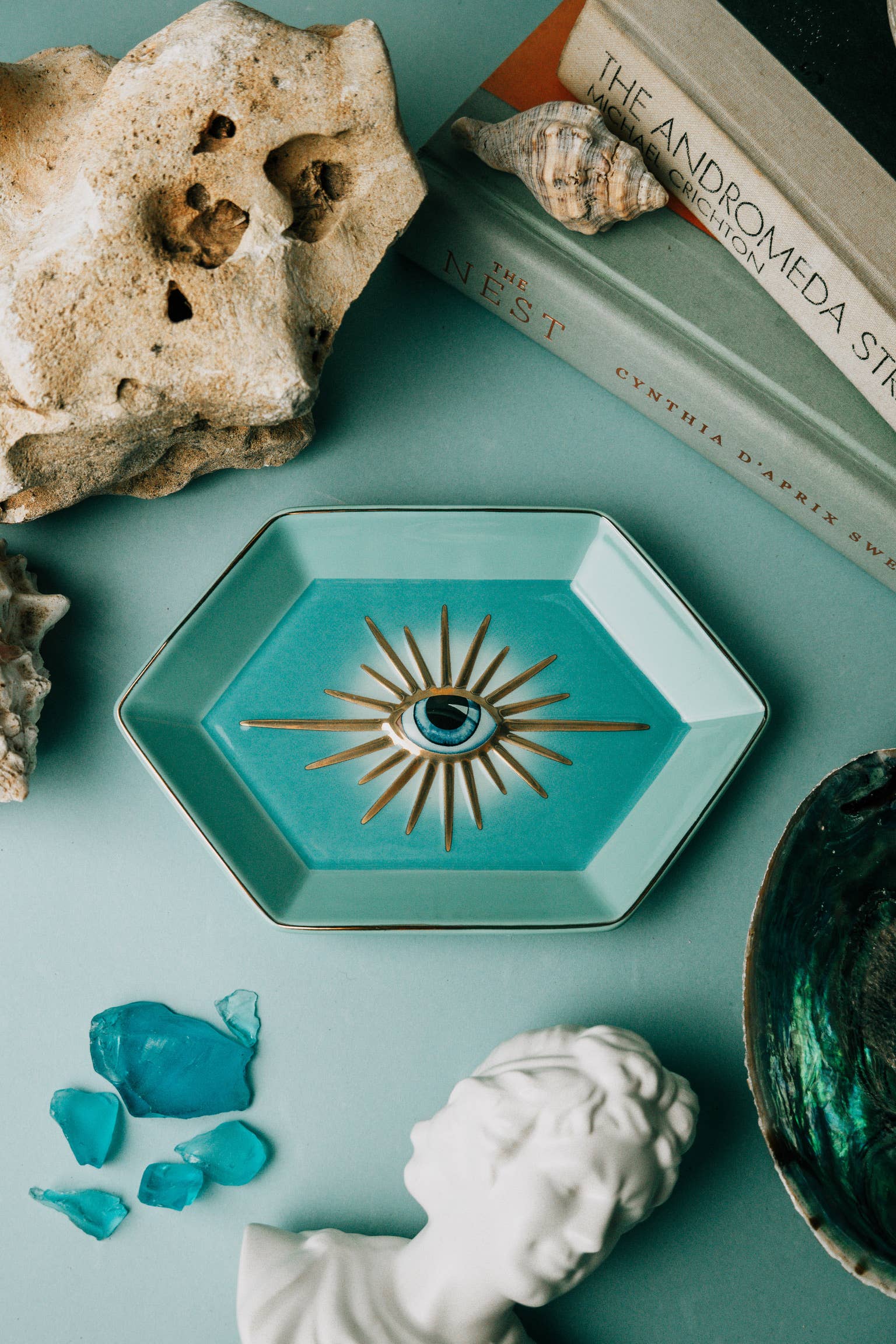 Teal decorative dish with eye design on a teal surface with rocks, books, and a bust.