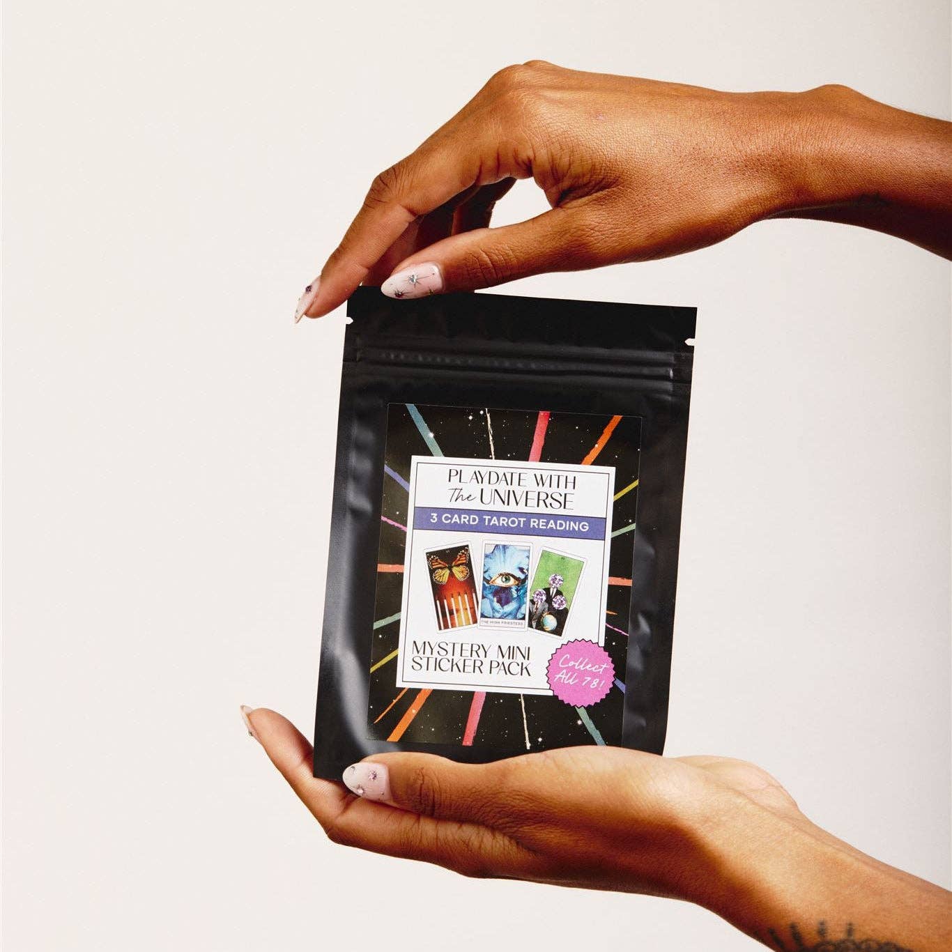 A person's hands holding a black resealable pouch package labeled 'Playdate with the Universe 3 Card Tarot Reading Mystery Mini Sticker Pack' against a plain light background.