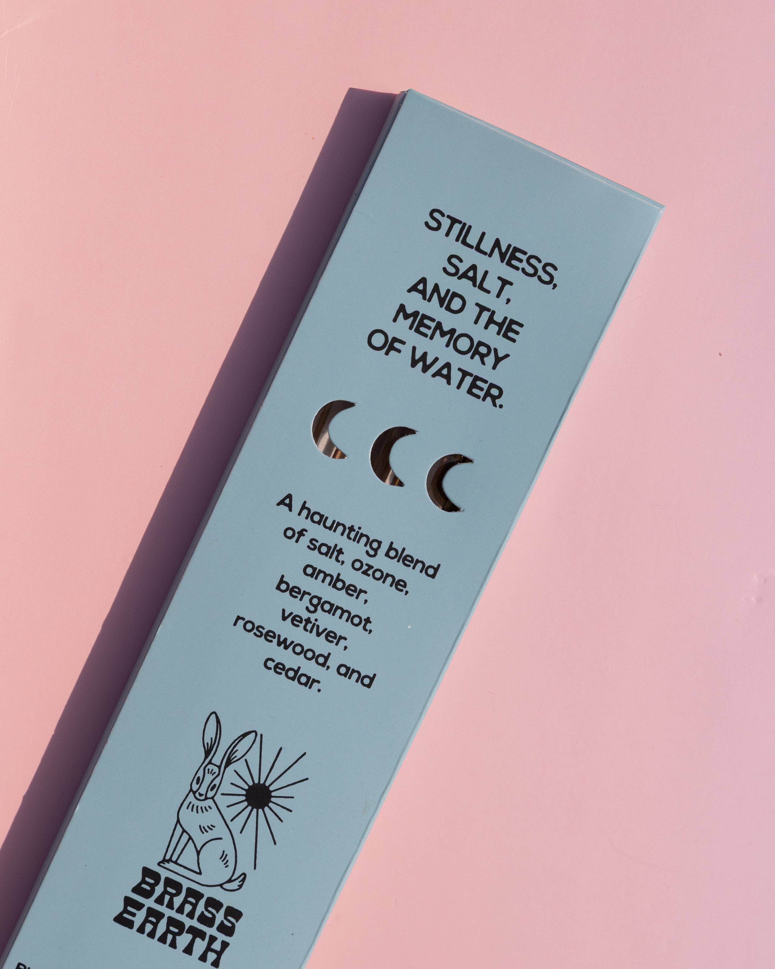 A light blue rectangular product box for Brass Earth incense sticks, photographed diagonally on a soft pink background. The box features black text describing a scent profile ('Stillness, Salt, and the Memory of Water') and a simple rabbit illustration.