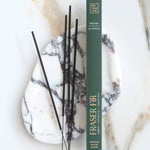 A vertical box of Botanica Fraser Fir incense sticks stands on a white marble tray with black incense sticks resting beside it on a light marble surface.