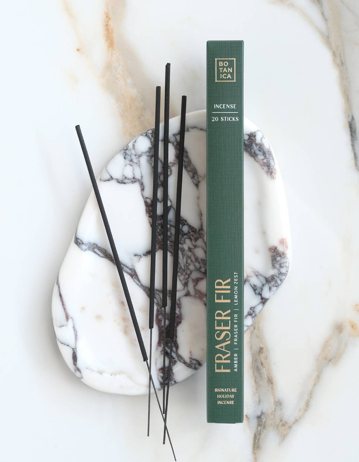 A vertical box of Botanica Fraser Fir incense sticks stands on a white marble tray with black incense sticks resting beside it on a light marble surface.