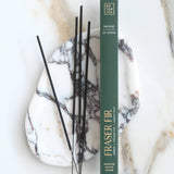 A vertical box of Botanica Fraser Fir incense sticks stands on a white marble tray with black incense sticks resting beside it on a light marble surface.