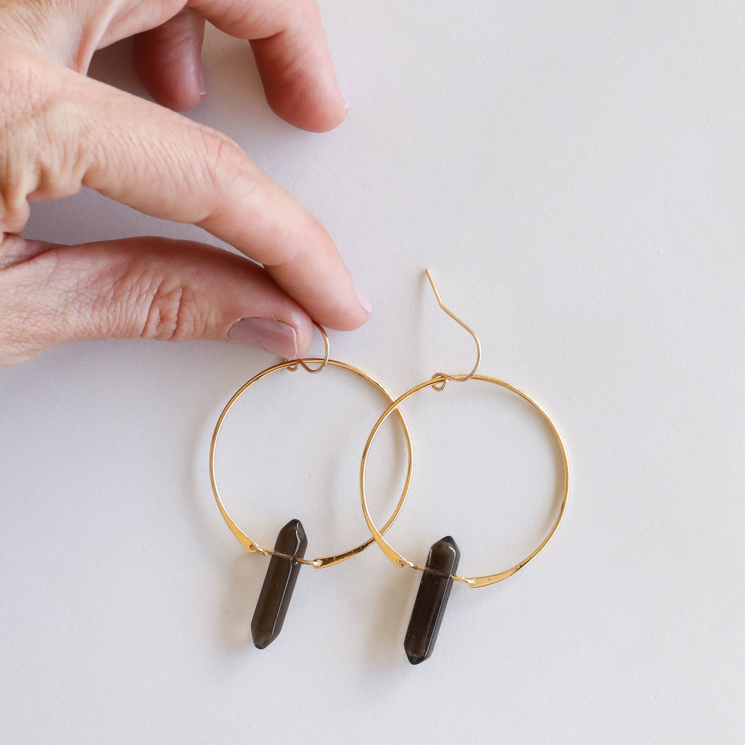 Gold hoop earrings with dark cylindrical beads held by a hand on a light background