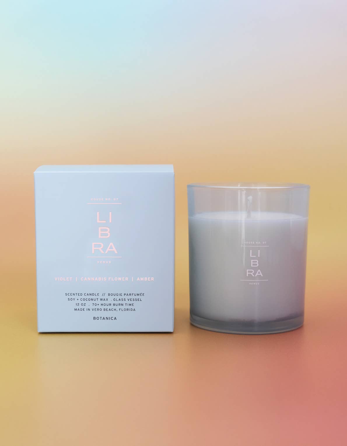 A scented candle in a frosted clear glass jar sits beside its light blue box on a smooth pastel gradient surface. The box and jar both show the brand name LIBRA and the scent name Violet + Cardamone Flower + Amber.