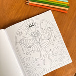 An open coloring book on a wooden table, showing a black-and-white line art page of a butterfly with the word 'AIR' at the top. Color pencils are lined up at the top of the frame.