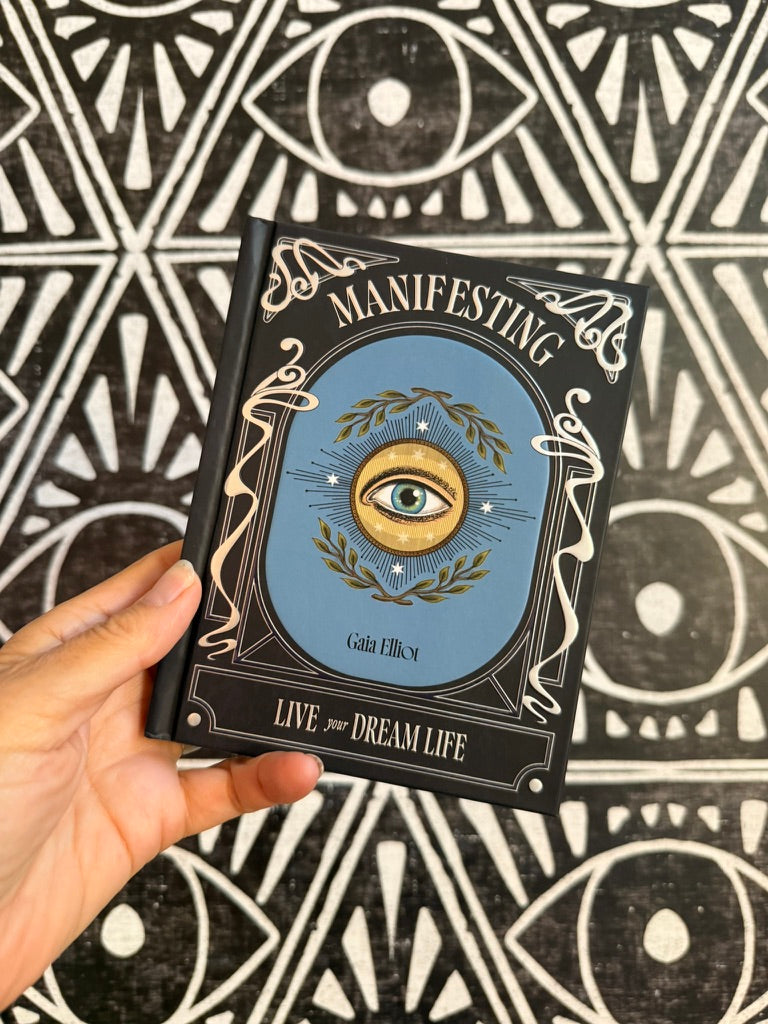 A hand holds a hardcover book titled 'Manifesting' by Gina Elliss, featuring a blue central illustration with an eye and gold laurel wreath, set against a black and white geometric patterned background.